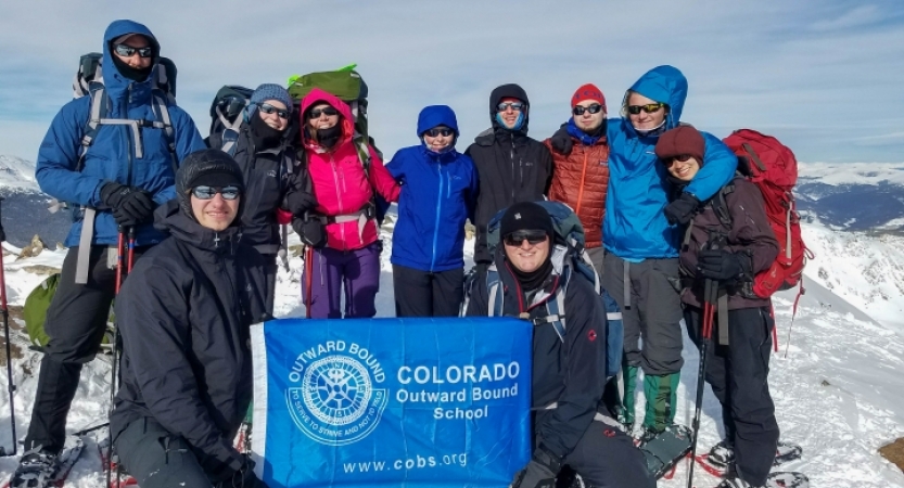 a group of people stand atop a snowy summit with an outward bound flag